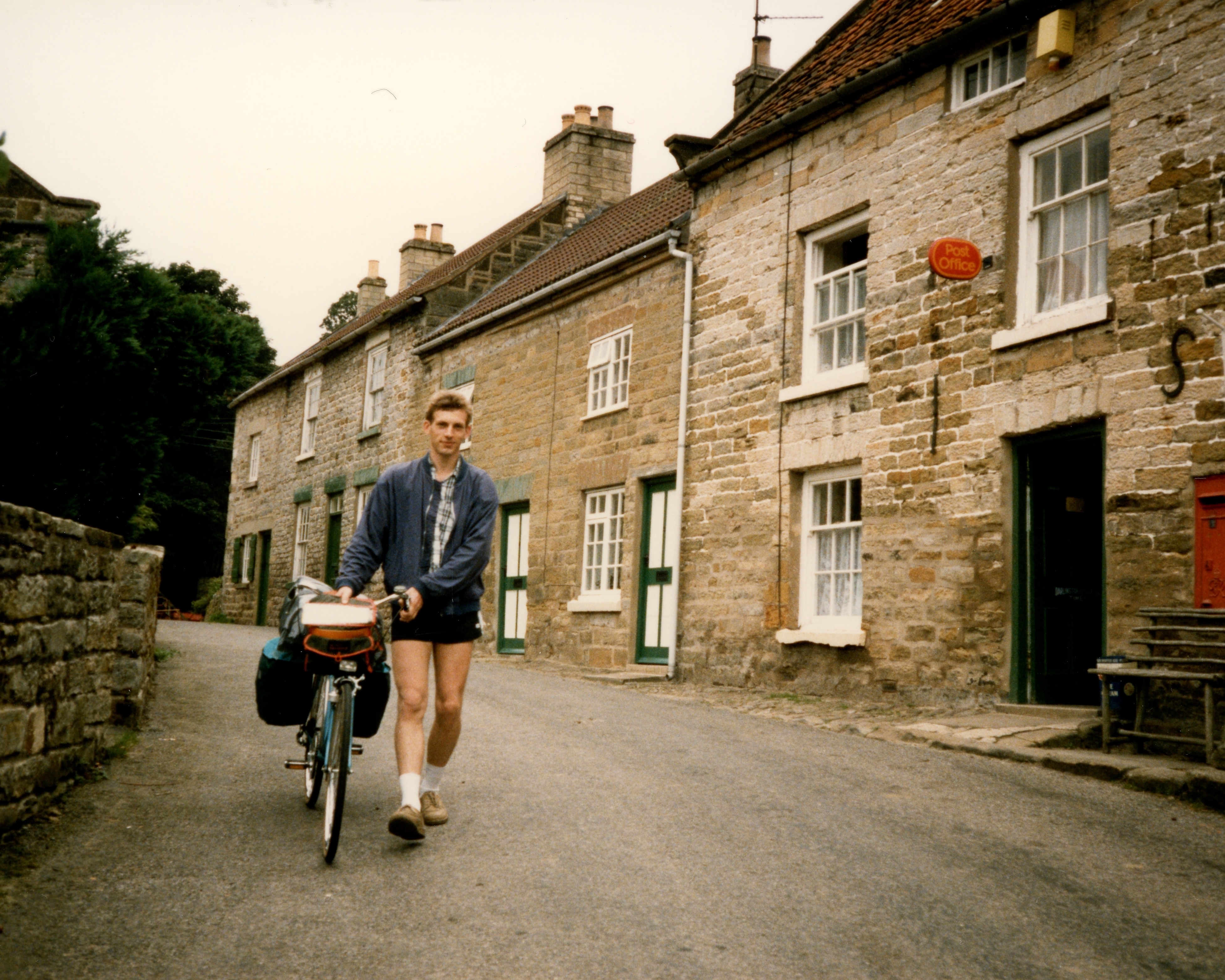 An old film photo showing a young man, my dad, walking through a Scottish village with his bike.